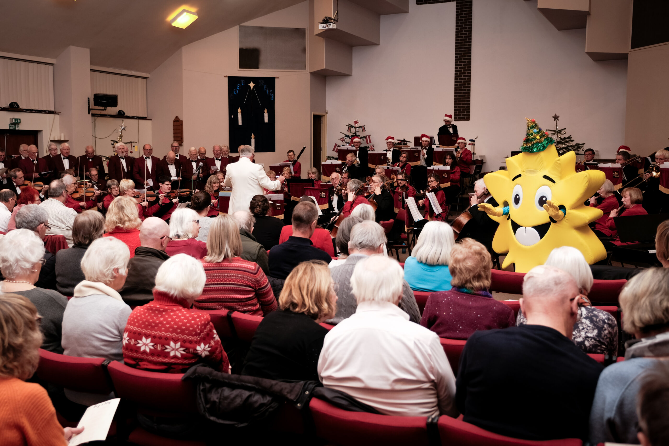 Seated audience watching Shrewsbury Light Orchestra perform with Lingen Davies Sunshine Mascot waving to the group.