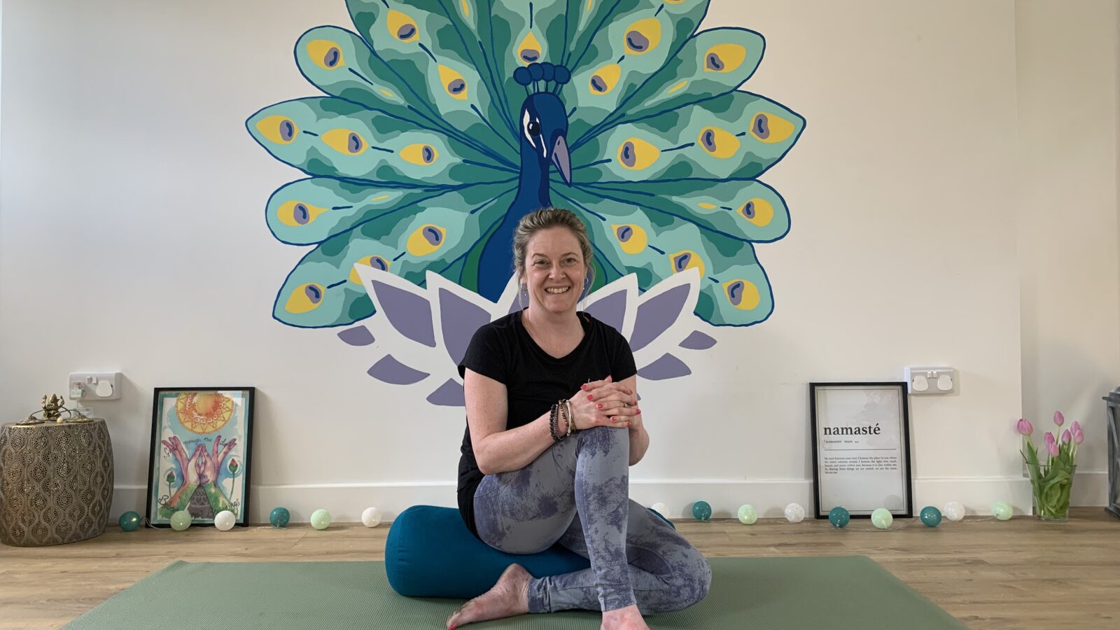 Smiling woman sits cross-legged on a yoga mat in front of a colorful wall mural of a peacock with open feathers. The serene studio space includes yoga props, framed artwork, a “Namasté” print, and decorative string lights along the floor. The woman wears comfortable activewear and appears ready for a mindfulness or yoga session, creating a peaceful and welcoming atmosphere.