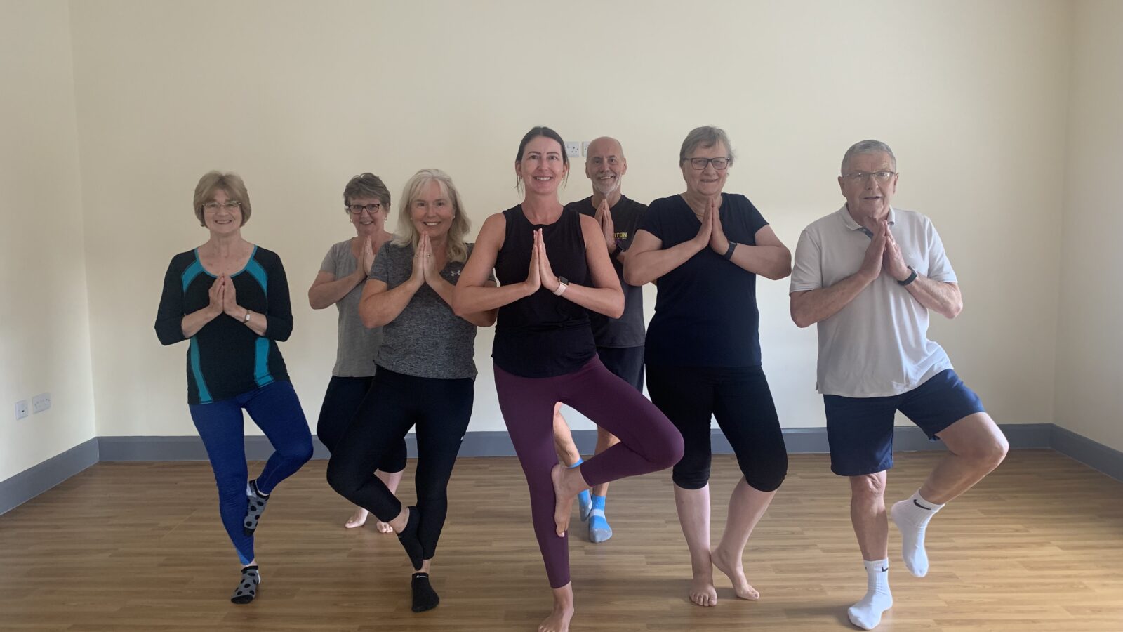 Group of seven people practicing yoga indoors, standing in the tree pose with palms together in a calm, light-filled studio. The participants, a mix of older adults and a younger instructor in the center, wear comfortable athletic clothing and smile as they balance on one leg. The wooden floor and neutral walls create a serene atmosphere, suggesting a community wellness or cancer recovery yoga class.