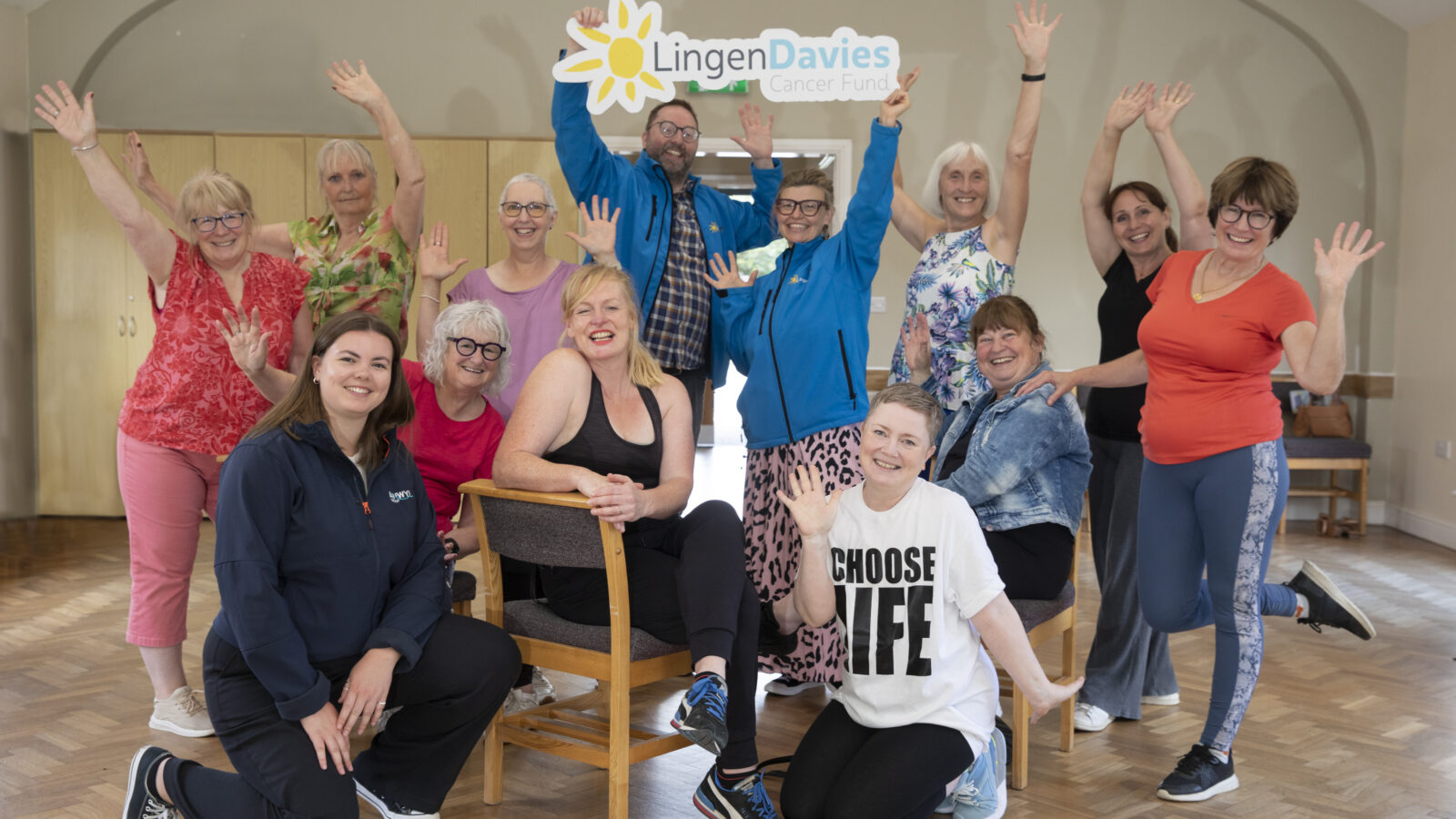Group of cheerful adults pose indoors, smiling and raising their hands beneath a large “Lingen Davies Cancer Fund” sign. The group, consisting mostly of women in colorful activewear, appears to be celebrating a community health or wellbeing session. The bright, welcoming room has wooden flooring and natural light, symbolizing positivity, cancer support, and active living.