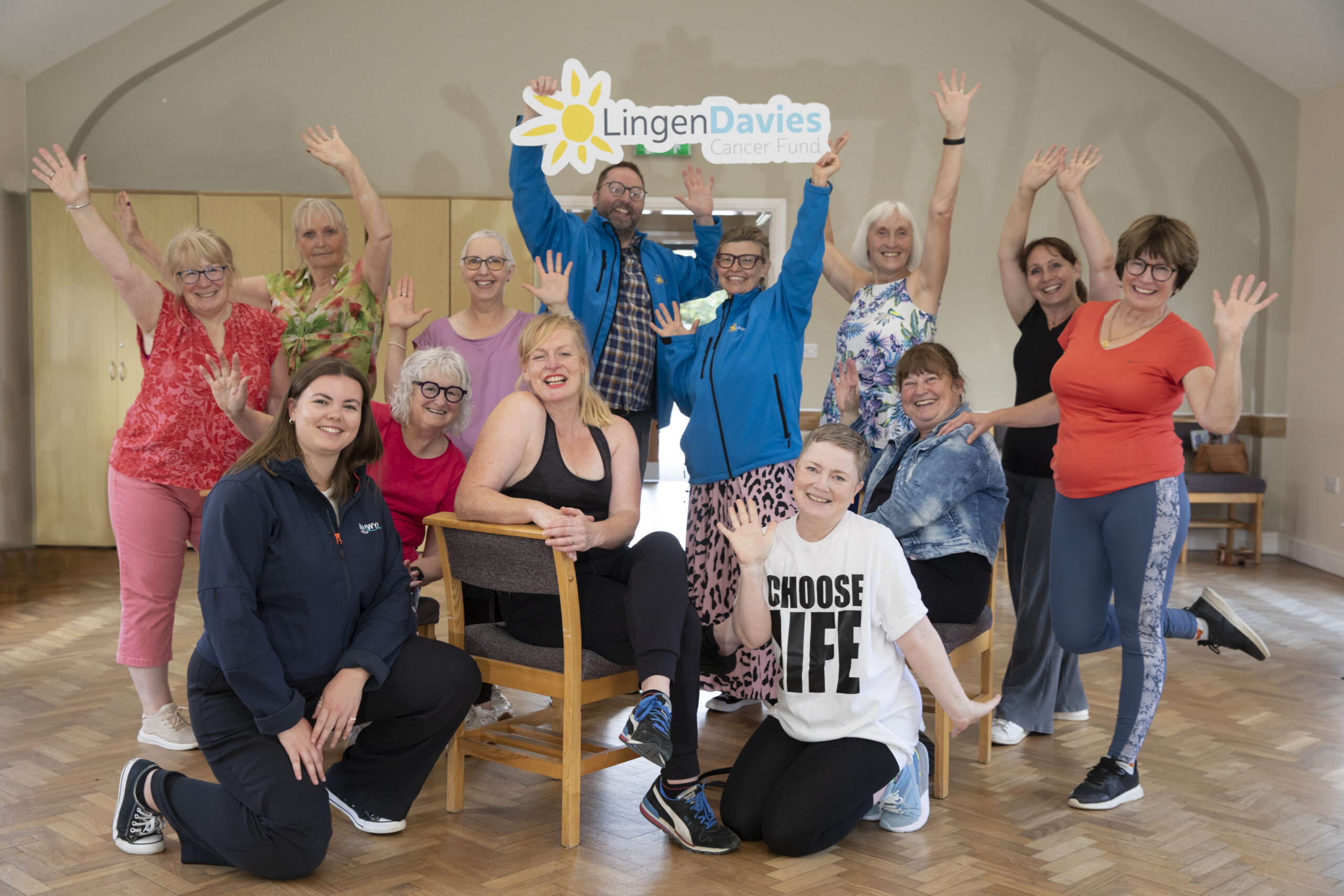Group of cheerful adults pose indoors, smiling and raising their hands beneath a large “Lingen Davies Cancer Fund” sign. The group, consisting mostly of women in colorful activewear, appears to be celebrating a community health or wellbeing session. The bright, welcoming room has wooden flooring and natural light, symbolizing positivity, cancer support, and active living.