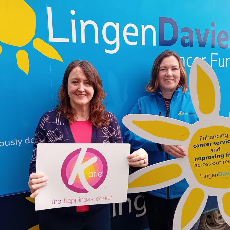 Two women stood in front of a blue background which reads, 'Lingen Davies Cancer Fund'. They are both holding large signs with logos.