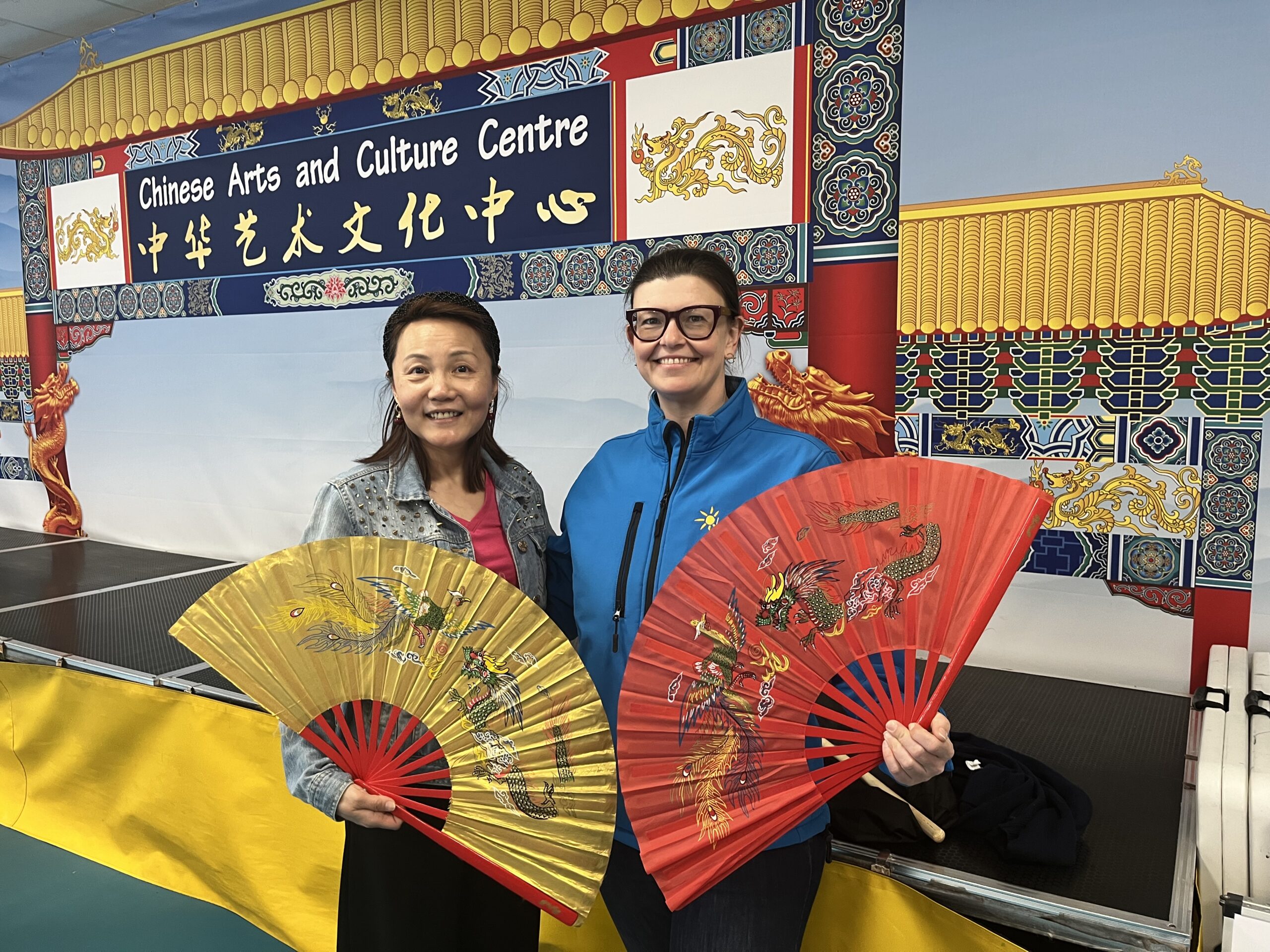 Two women posing for a camera. They are both holding open ornate fans.