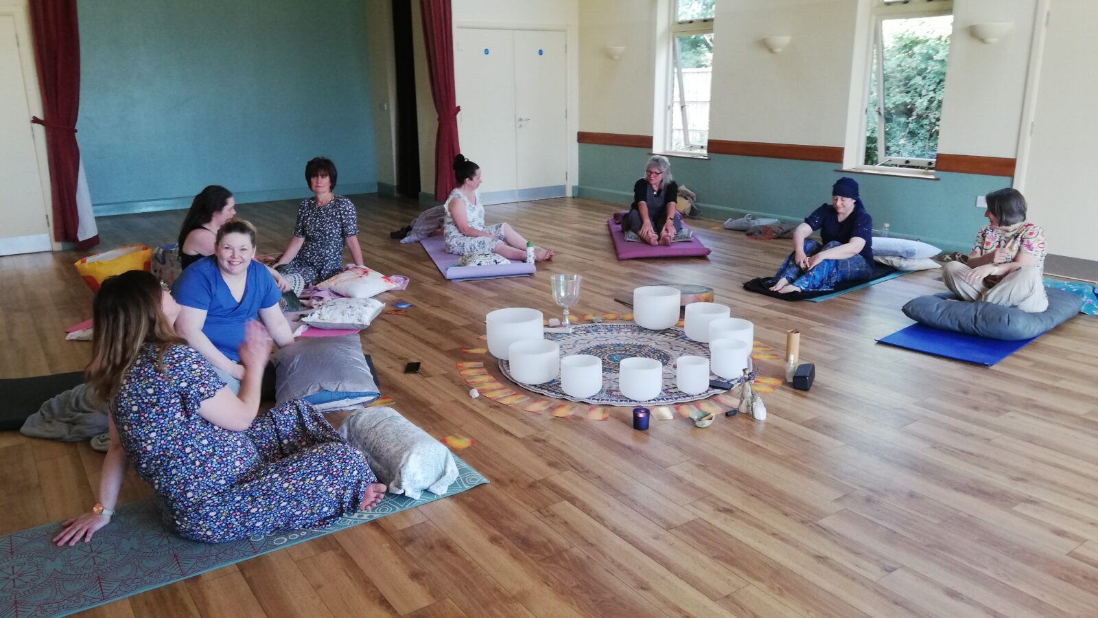 Group of women sitting on yoga mats and cushions in a calm, softly lit room during a relaxation or sound healing session. The participants form a circle around a central display of crystal singing bowls, candles, and decorative cloth, creating a peaceful, meditative atmosphere. The wooden floor and natural light enhance the sense of tranquility.