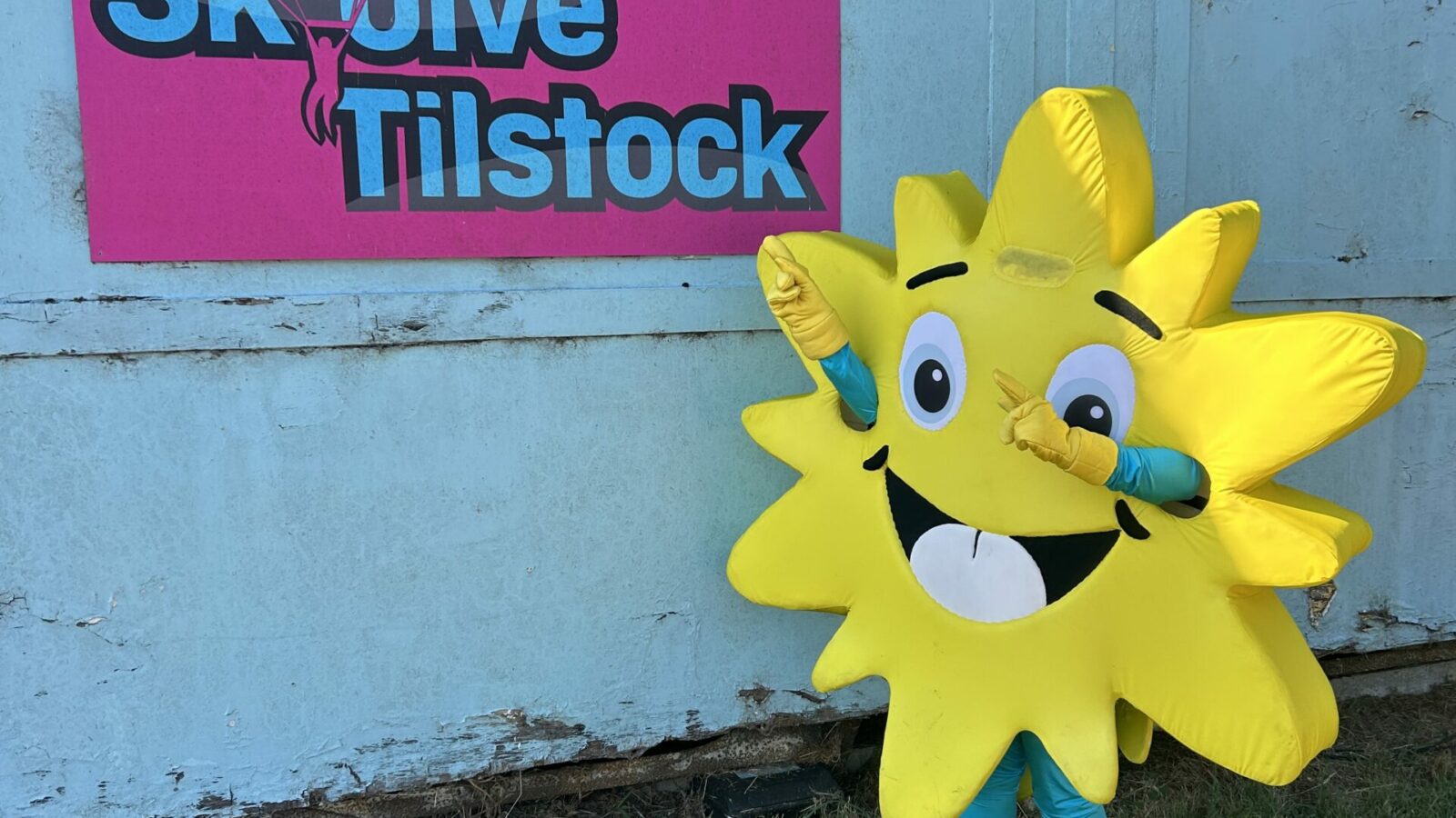 Person wearing a bright yellow sun mascot costume poses cheerfully beside a colorful “Skydive Tilstock” sign on a blue wooden wall. The mascot, with a smiling face and cartoon eyes, stands on a grassy area under clear sunlight, promoting a skydiving charity event linked to the Lingen Davies Cancer Support.