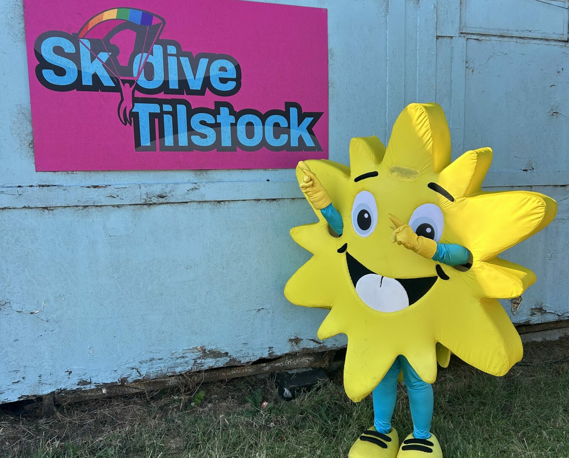 Person wearing a bright yellow sun mascot costume poses cheerfully beside a colorful “Skydive Tilstock” sign on a blue wooden wall. The mascot, with a smiling face and cartoon eyes, stands on a grassy area under clear sunlight, promoting a skydiving charity event linked to the Lingen Davies Cancer Support.