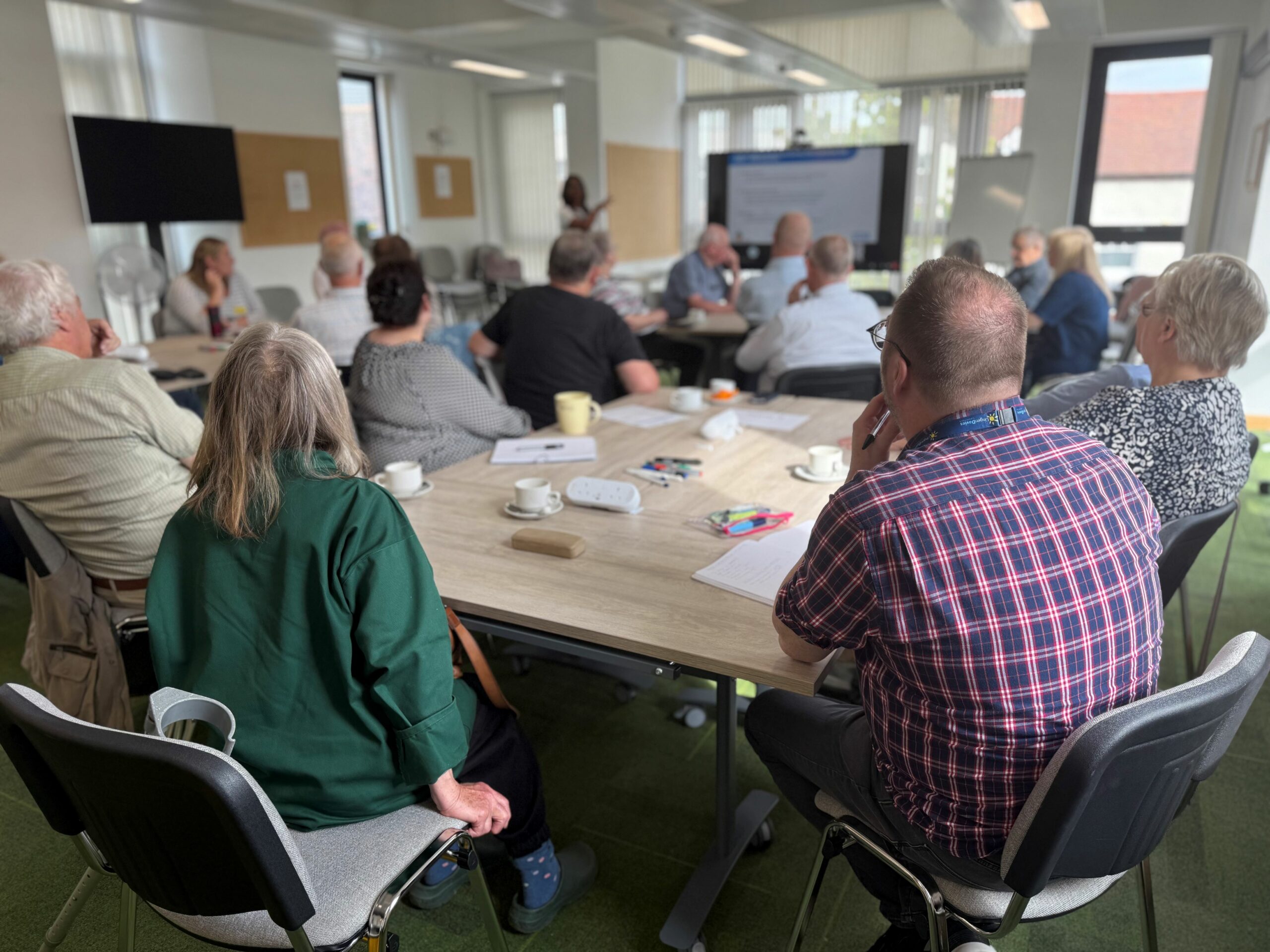 A group of people sits around tables in a bright meeting room, attentively listening to a presenter at the front near a large screen. Papers, notebooks, and coffee cups are scattered on the tables, creating a casual, collaborative atmosphere.