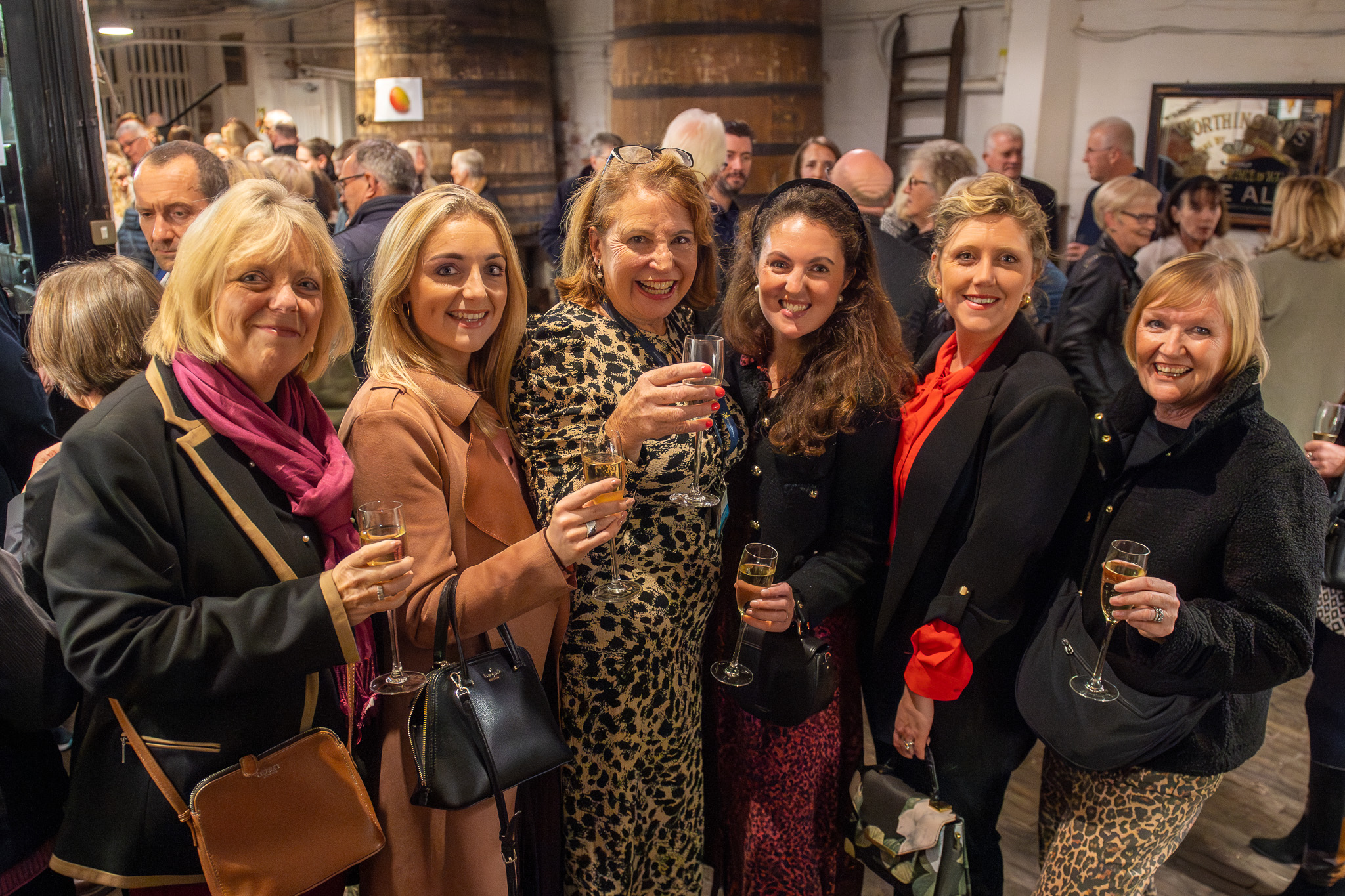 A group of six smiling women stand close together at a crowded indoor event, each holding a glass of sparkling wine and posing for the camera.