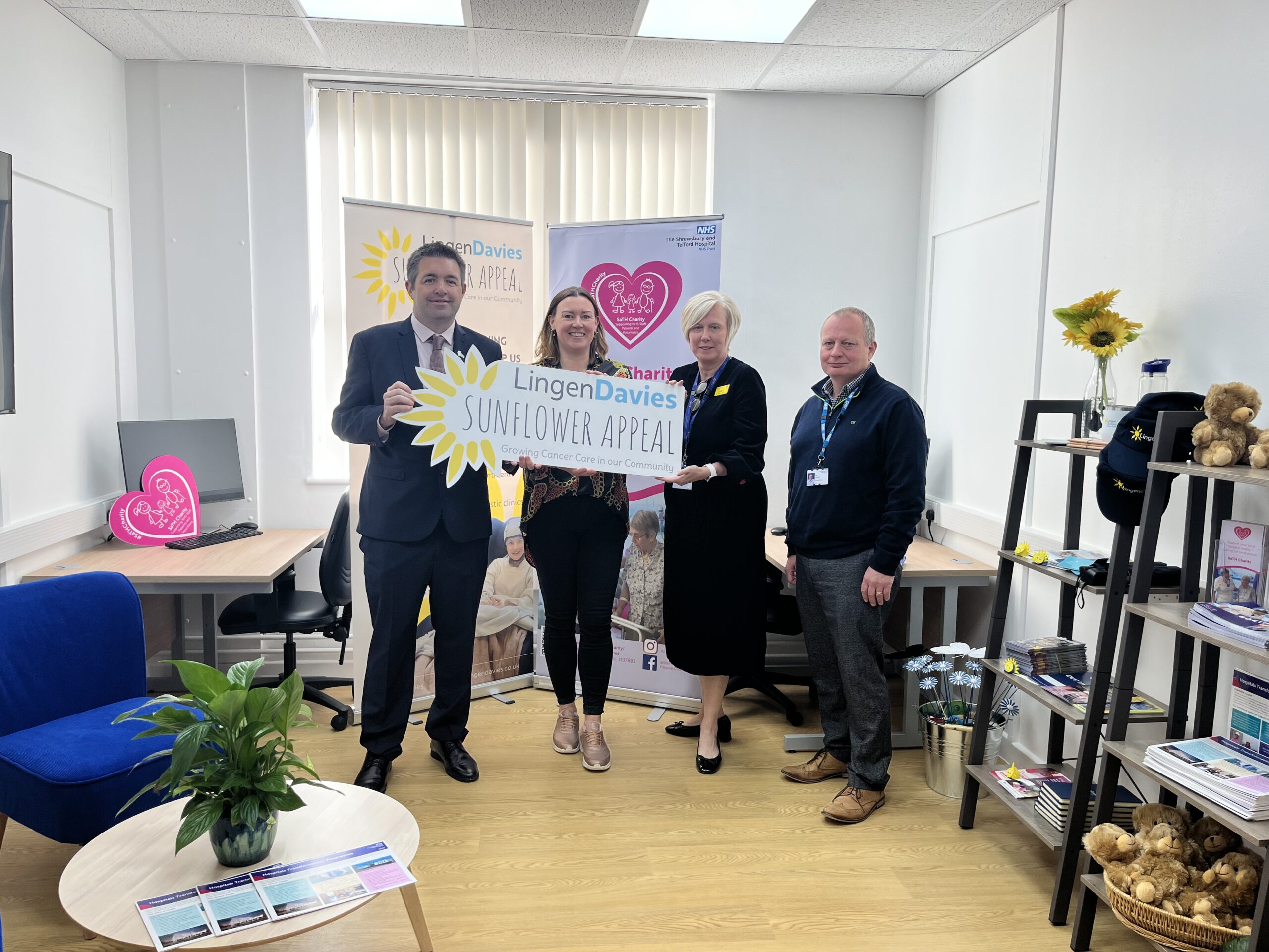 Four people stand together in an office holding a large Lingen Davies Sunflower Appeal sign, smiling in front of promotional banners and sunflower decorations.