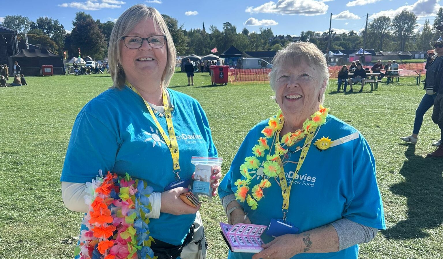 Two smiling women wearing blue Lingen Davies Cancer Support T-shirts stand outdoors on a sunny day, holding raffle tickets and donation tins at Heal Festival.