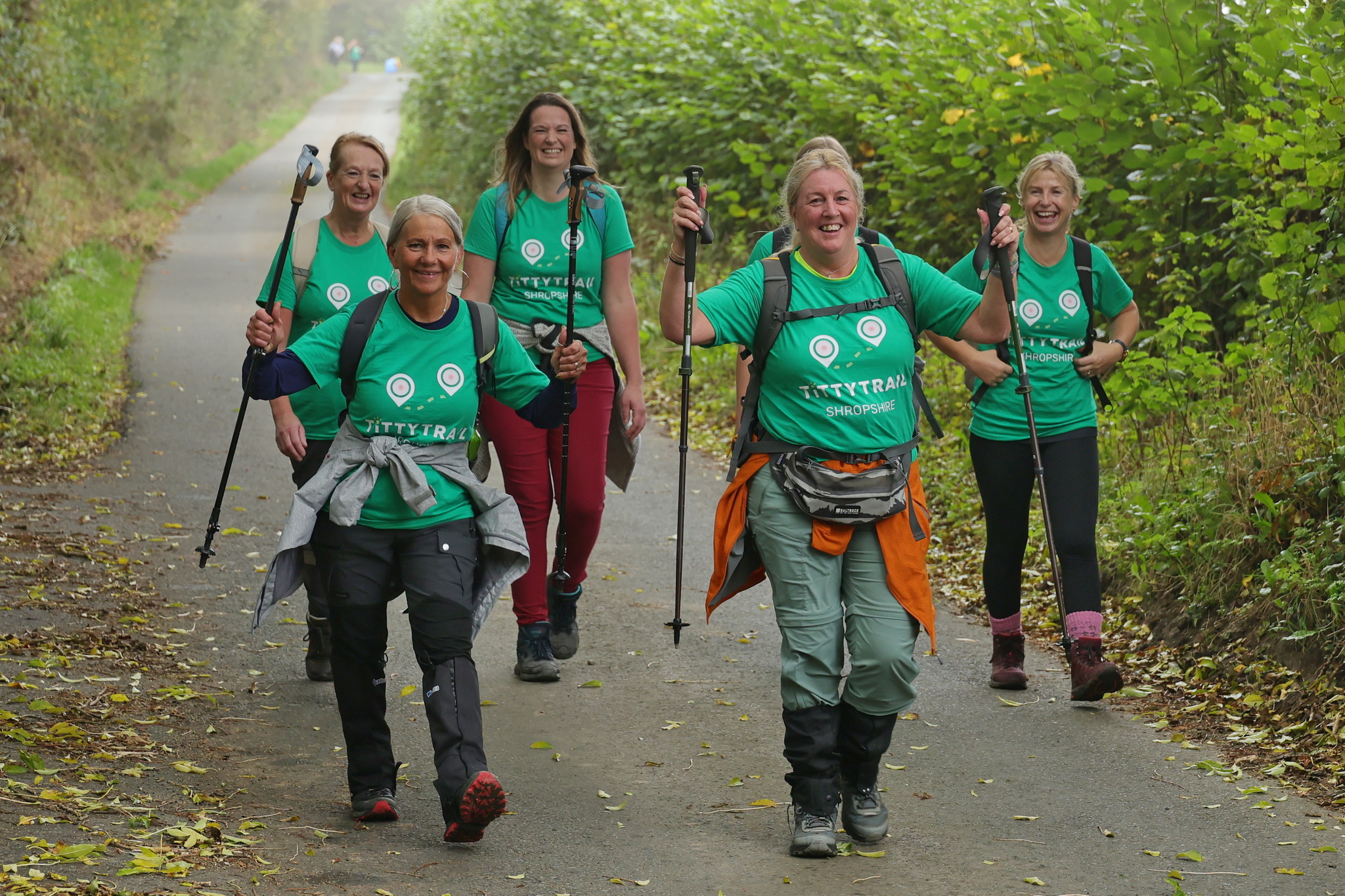A group of five smiling women in matching green “Titty Trail Shropshire” T-shirts walk along a leafy country lane, using walking poles during a charity hike.
