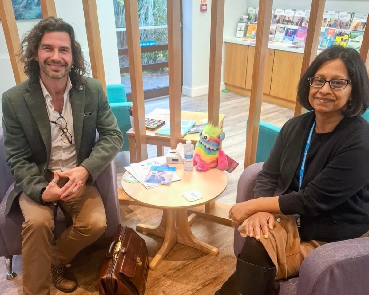 A man and a woman sit facing each other in a bright, welcoming lounge with pamphlets on display behind them. A rainbow-colored stuffed creature sits on the table between them.