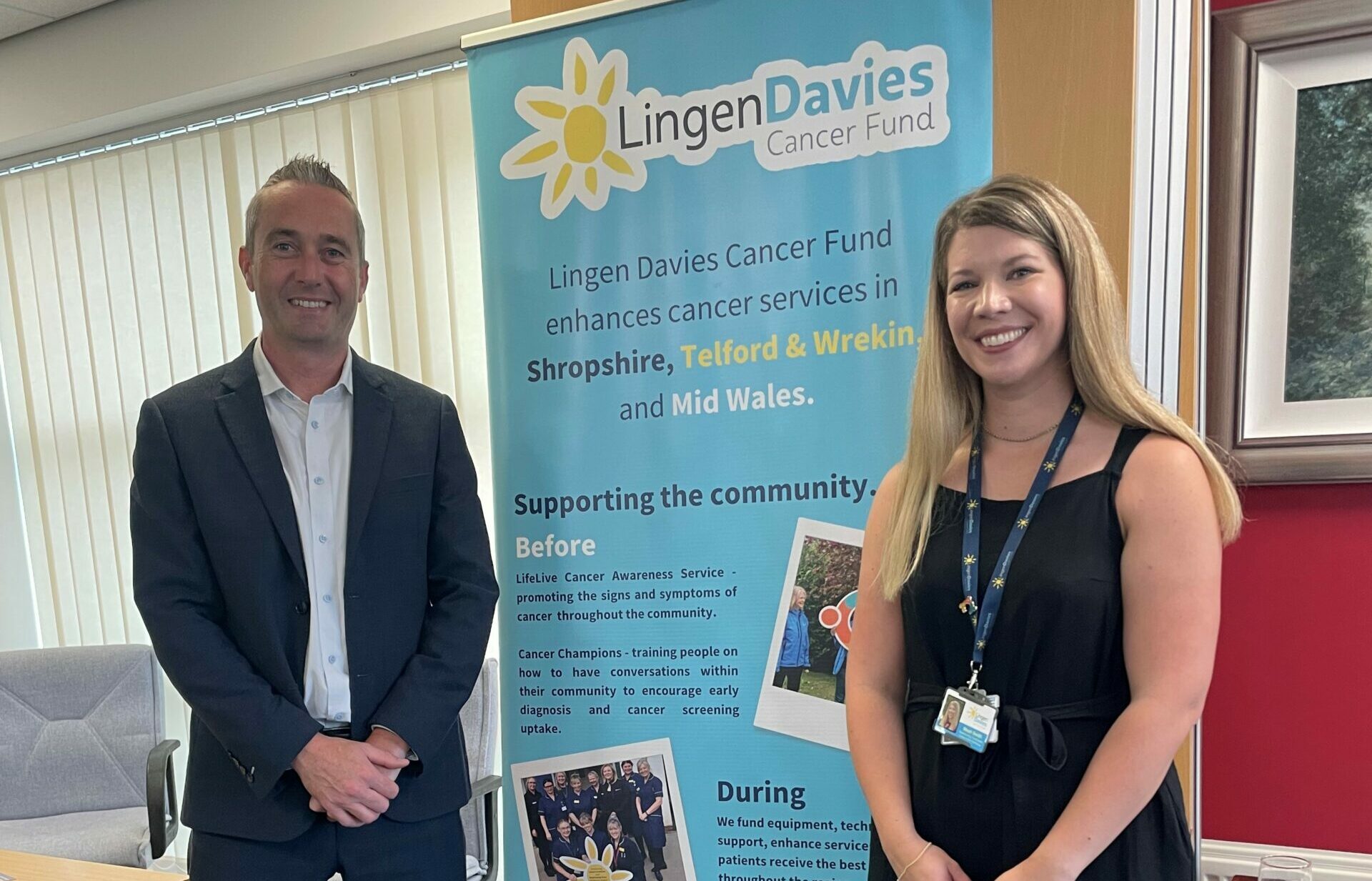 Two people pose in front of a Lingen Davies Cancer Fund display that highlights community cancer awareness and support. Both are smiling in a bright office setting.