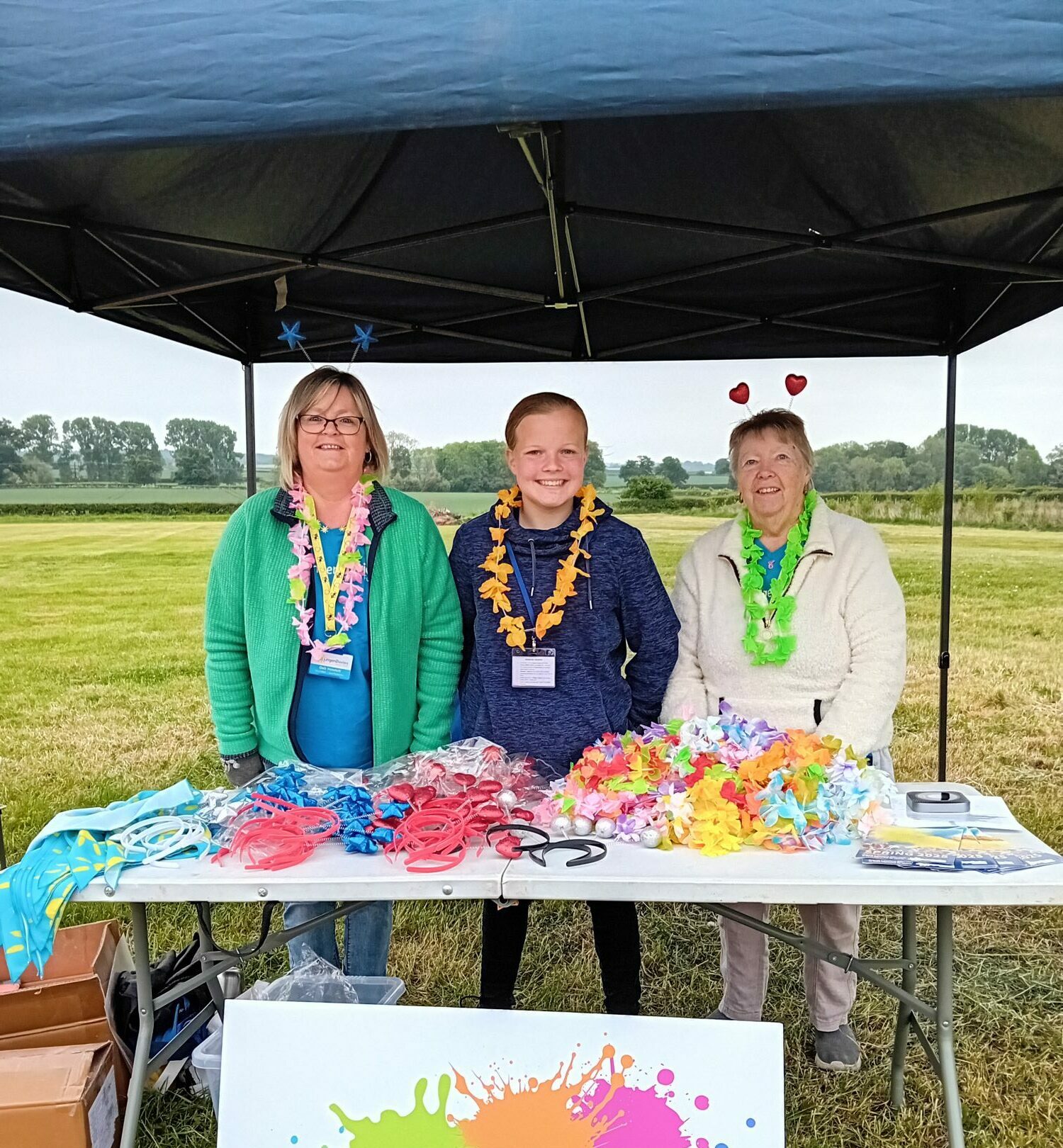 Outdoor Colour Fest 5K charity event stall with three smiling volunteers standing under a black canopy tent, each wearing colorful Hawaiian leis and fun headbands; table displays bright flower garlands, neon headbands, accessories, registration materials, and donation items. Large sign in front shows multicolored paint splashes with text
