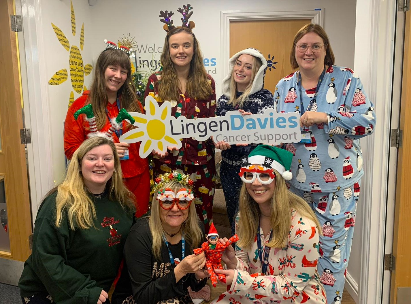 Seven smiley women in Christmas pyjamas and festive headwear hold a Lingen Davies Cancer Support sign