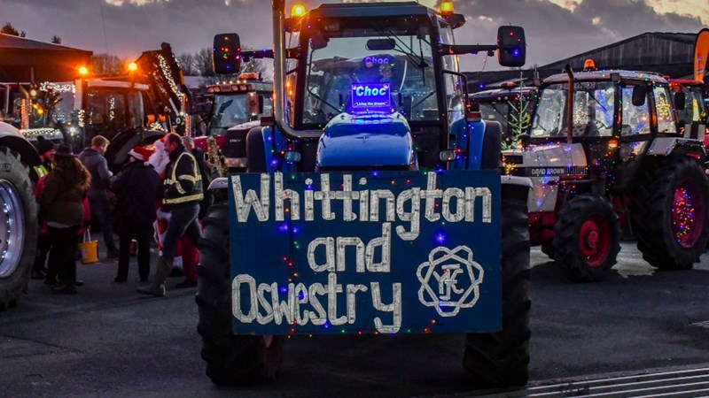 A blue tractor decorated with festive lights and a large sign on the front reading ‘Whittington and Oswestry YFC’ in glittery letters, with the Young Farmers Club logo. The tractor is part of a lineup of illuminated tractors, all adorned with colourful lights, parked in an outdoor area at dusk. Several people in winter clothing are gathered nearby, some wearing Santa hats, adding to the festive atmosphere.