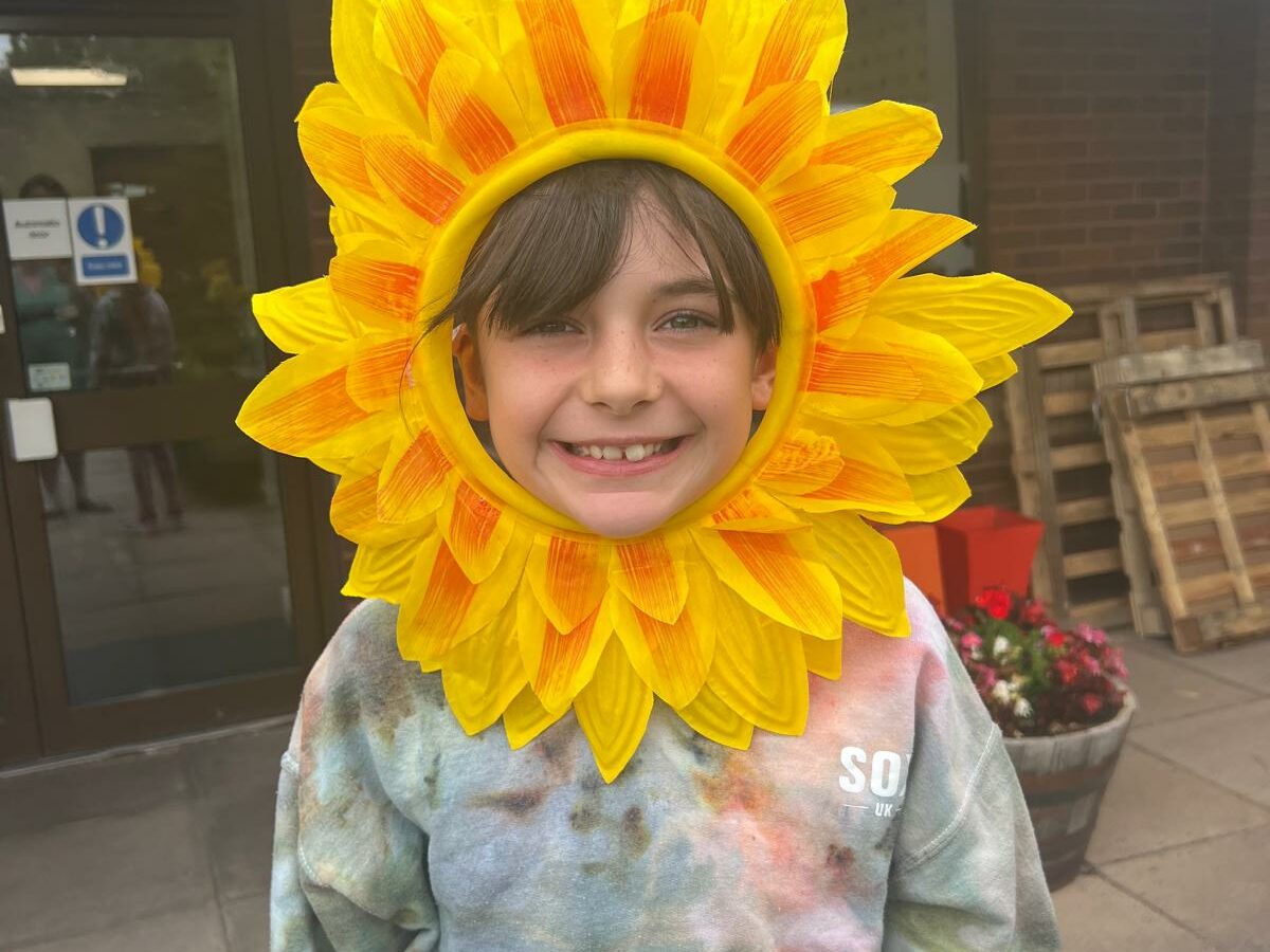 A person wearing a large sunflower headpiece stands outside a brick building near a fundraising office entrance. They are dressed in a multicoloured tie‑dye hoodie, and there are wooden pallets and a planter of red and white flowers in the background.